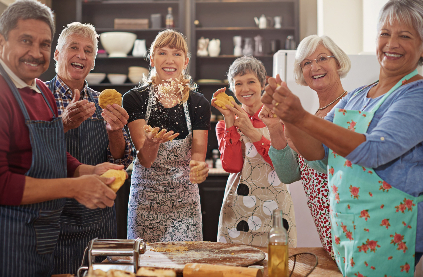 A group of older adults in a baking class with their instructor, each holding dough, smiling and looking directly at the camera
