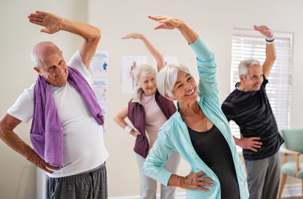 A group of older adults in an exercise class smiling and stretching with one hand over their heads