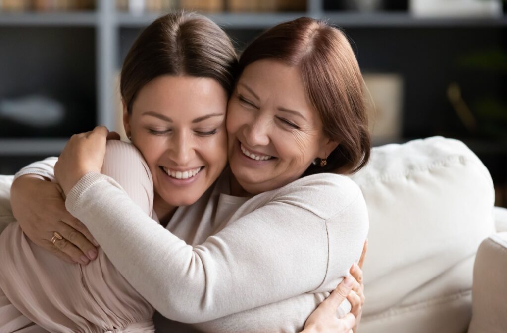 An older adult woman with her daughter sitting on the couch, smiling and hugging each other.