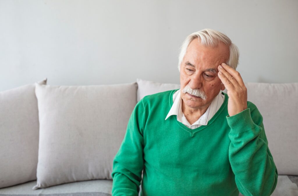 Senior man sitting on the couch with his hand to his head looking upset and uncomfortable.